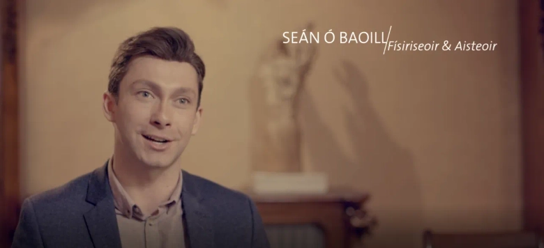 Smiling man in a blazer with text overlay "Seán Ó Baoill, Físiriseoir & Aisteoir" against a blurred interior background.